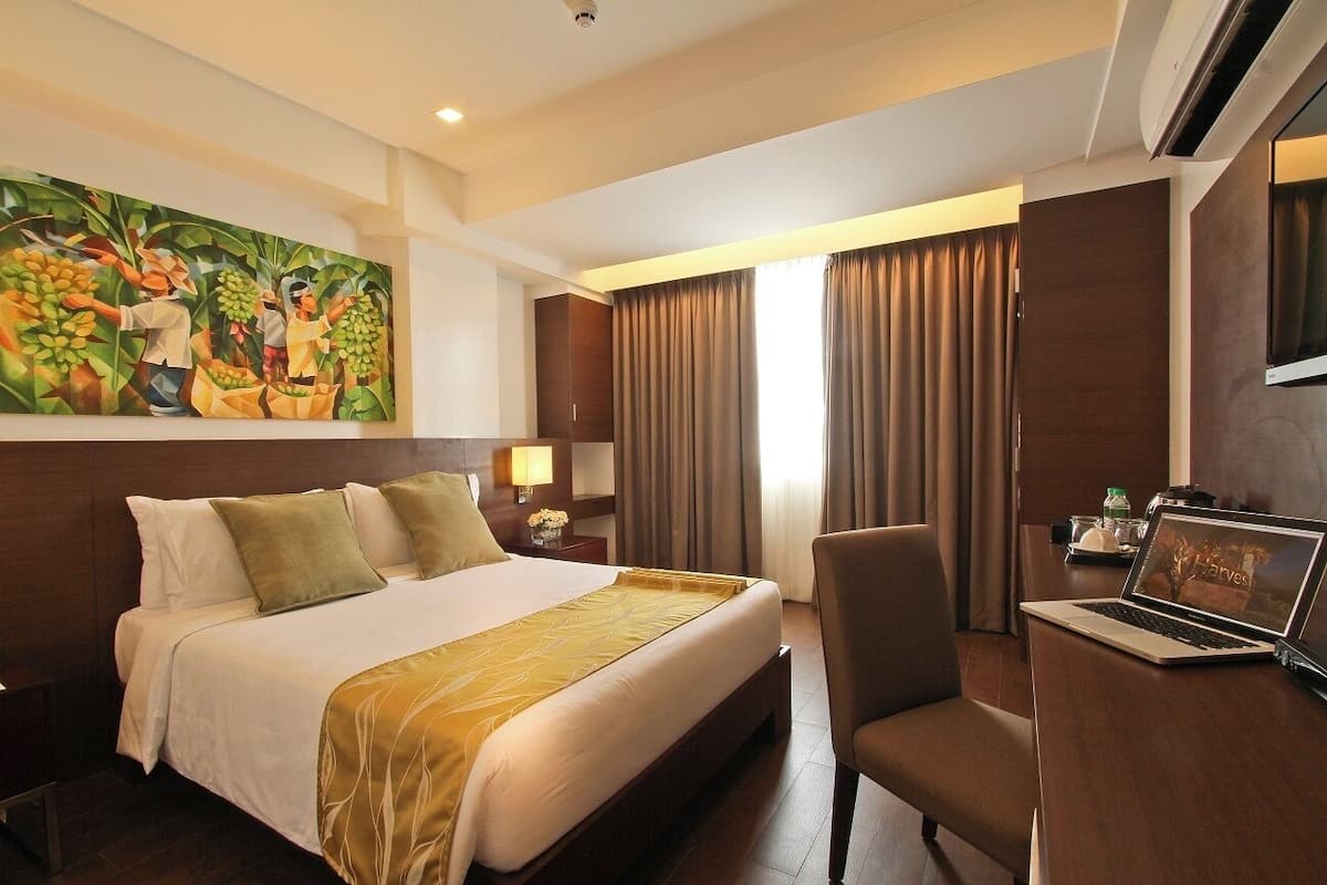 A hotel room with a large bed, a desk with a laptop, a painting on the wall, and a window with curtains.