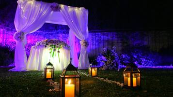 An outdoor wedding arch with white drapery and floral arrangements, illuminated by lanterns and colored lights.