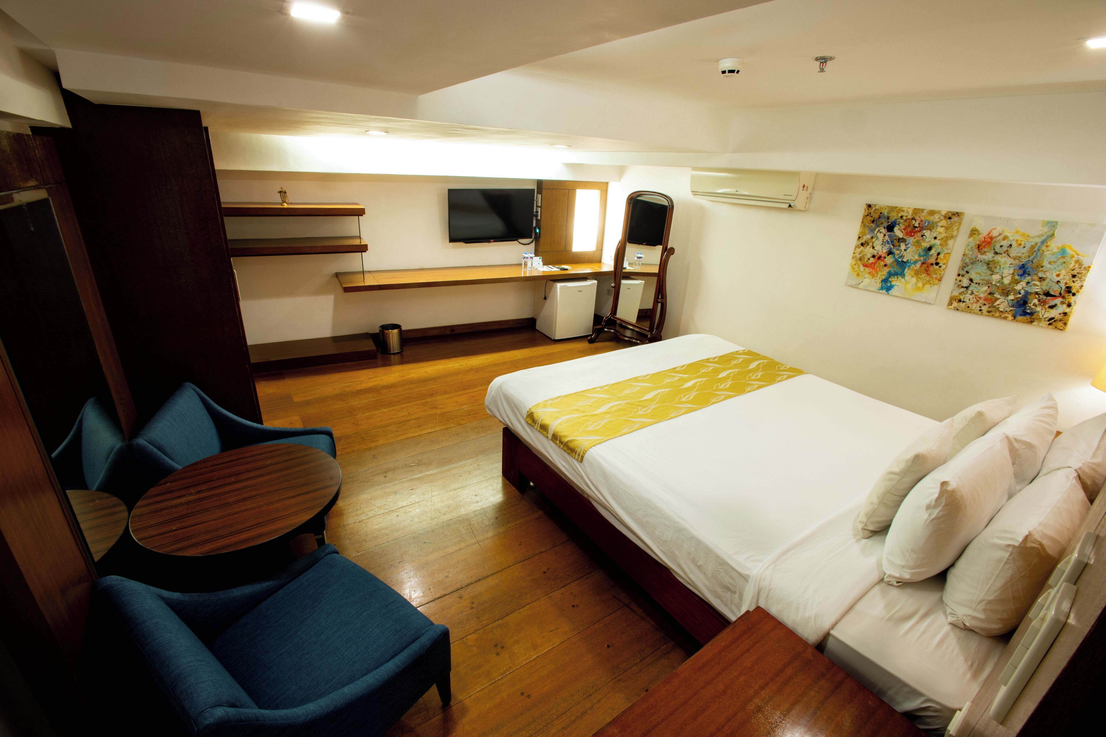 A hotel room with a bed, a desk with a chair, a small refrigerator, a television, and artwork on the wall.