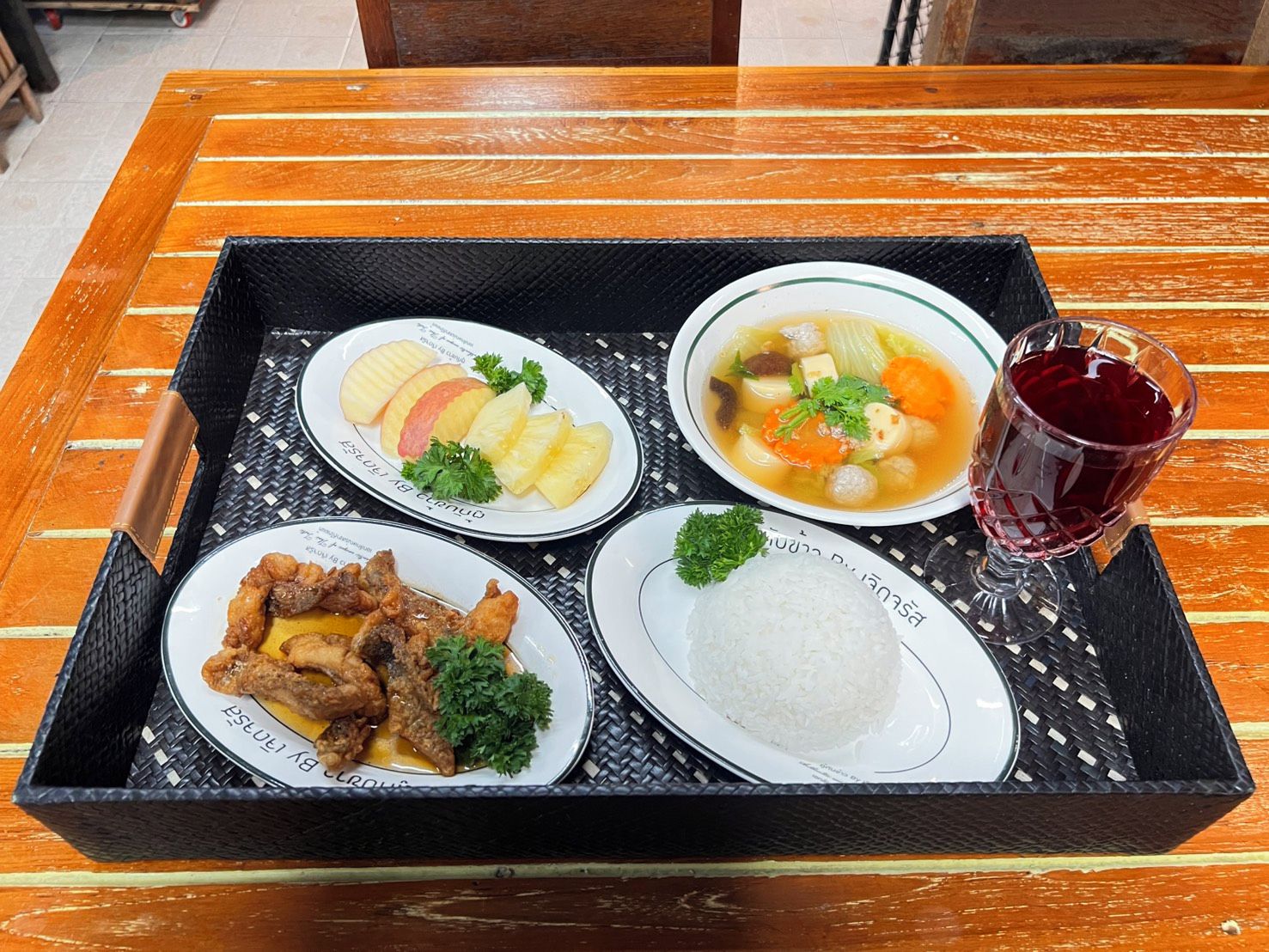 Un plateau contenant une variété de plats, notamment un bol de soupe, une assiette de viande, une assiette de fruits et un verre d’une boisson rouge.