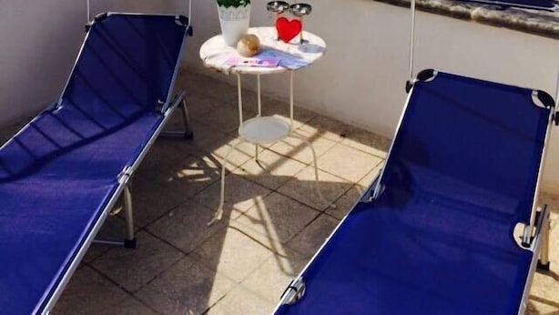 A balcony with two blue sun loungers, a small round table with a potted plant, a red heart-shaped decoration, and a view of the sea. - B&B Petali Rosa (Polignano a Mare)