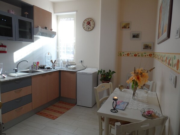A kitchen with a sink, stove, and cabinets. There is a window with curtains, a clock on the wall, and a table with chairs. - B&B Petali Rosa (Polignano a Mare)