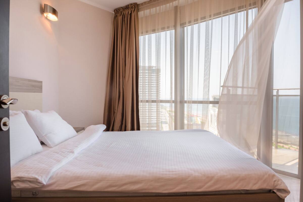 A hotel room with a large bed, a view of the city through sheer curtains, and a balcony with a view of the sea.