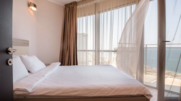 Habitación de hotel con una cama grande, vistas de la ciudad a través de cortinas translúcidas y un balcón con vistas al mar.