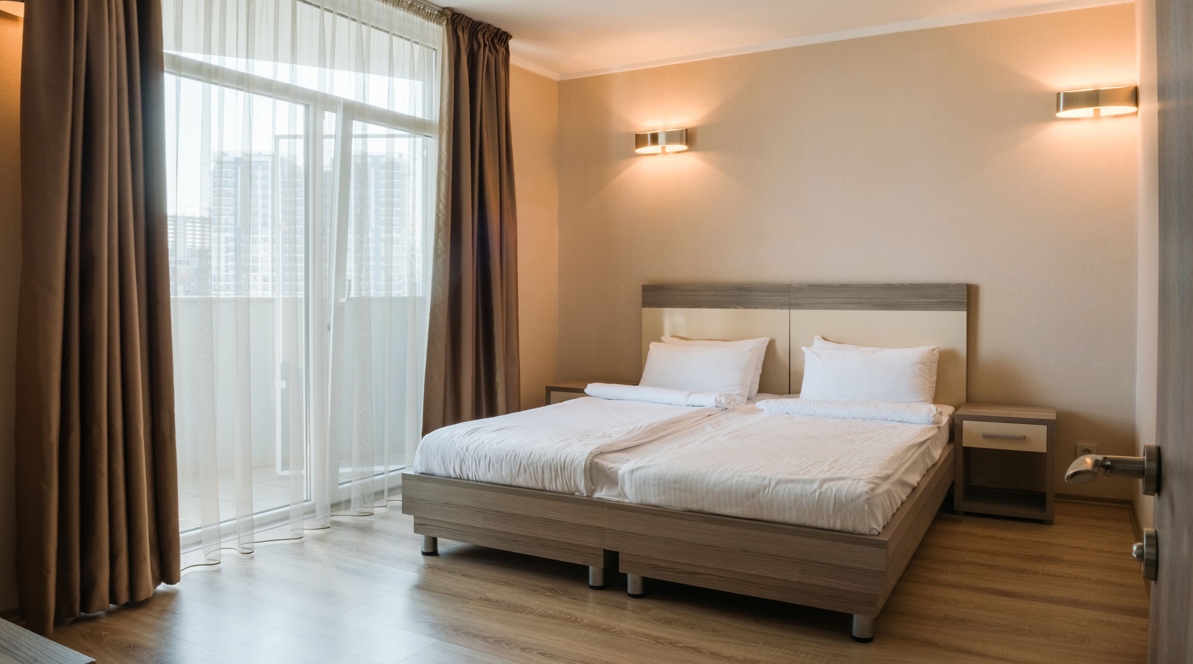 A hotel room with a large bed, two bedside tables, a wooden headboard, and a view of buildings through sheer curtains.