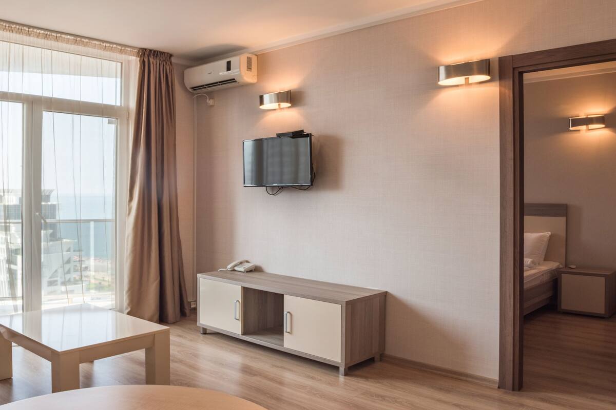 A modern hotel room with a large window, a flat-screen TV mounted on the wall, a bedside table with a phone, and a separate sleeping area with a bed and nightstand.