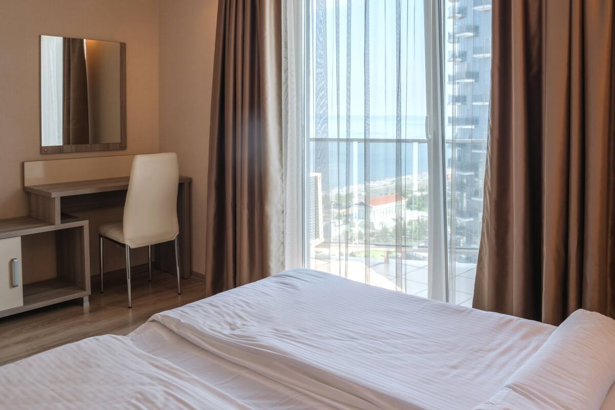 A hotel room with a large bed, a desk, a chair, and a view of a cityscape through large windows.