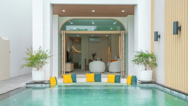 Un área de piscina interior moderna con vista despejada hacia la habitación contigua, que incluye un espacio de descanso con cojines y plantas en macetas.
