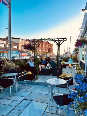 An outdoor seating area with tables and chairs, a pergola with plants, and a view of the street with buildings and shops. - Danescourt Lodge (Blackpool)