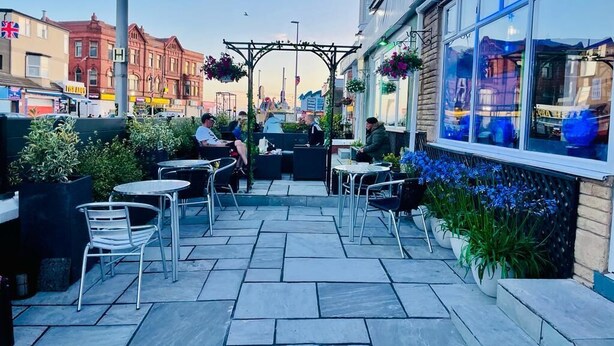 Un patio al aire libre con mesas y sillas, rodeado de macetas con plantas y flores.