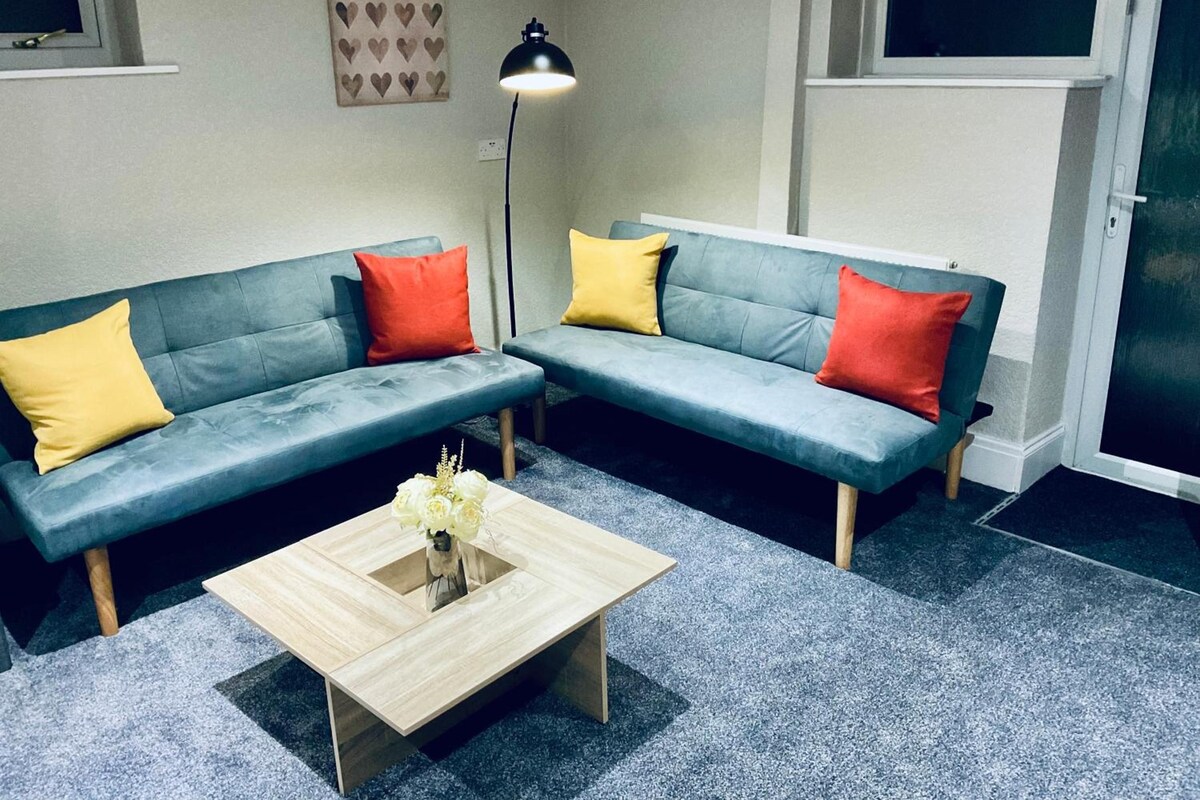 Una sala de estar moderna con dos sofás grises, uno con un cojín amarillo y el otro con uno rojo, una mesa de centro de madera con un jarrón de flores y una lámpara de pie.