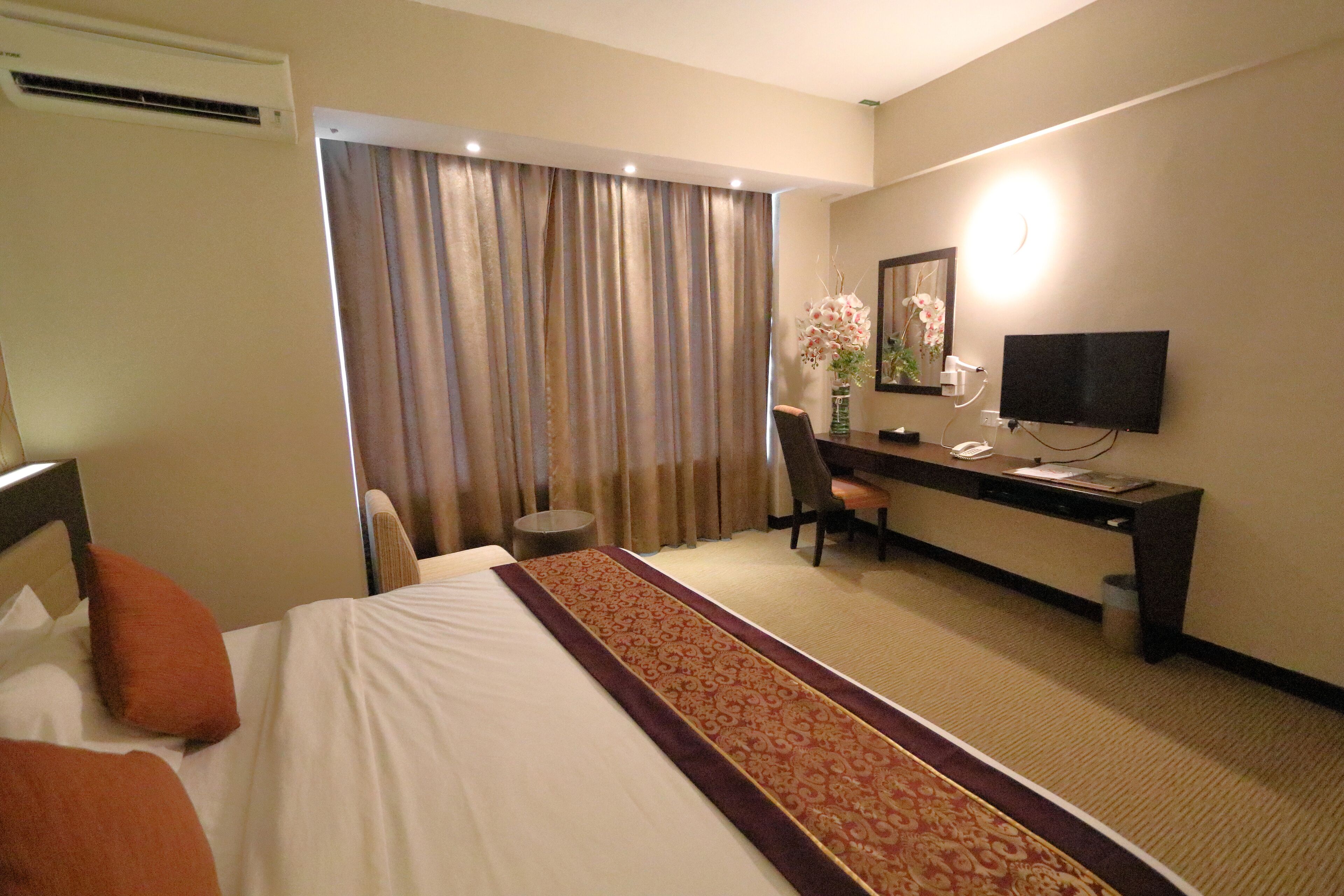 Habitación de hotel con cama, escritorio, silla, televisor y ventana con cortinas.