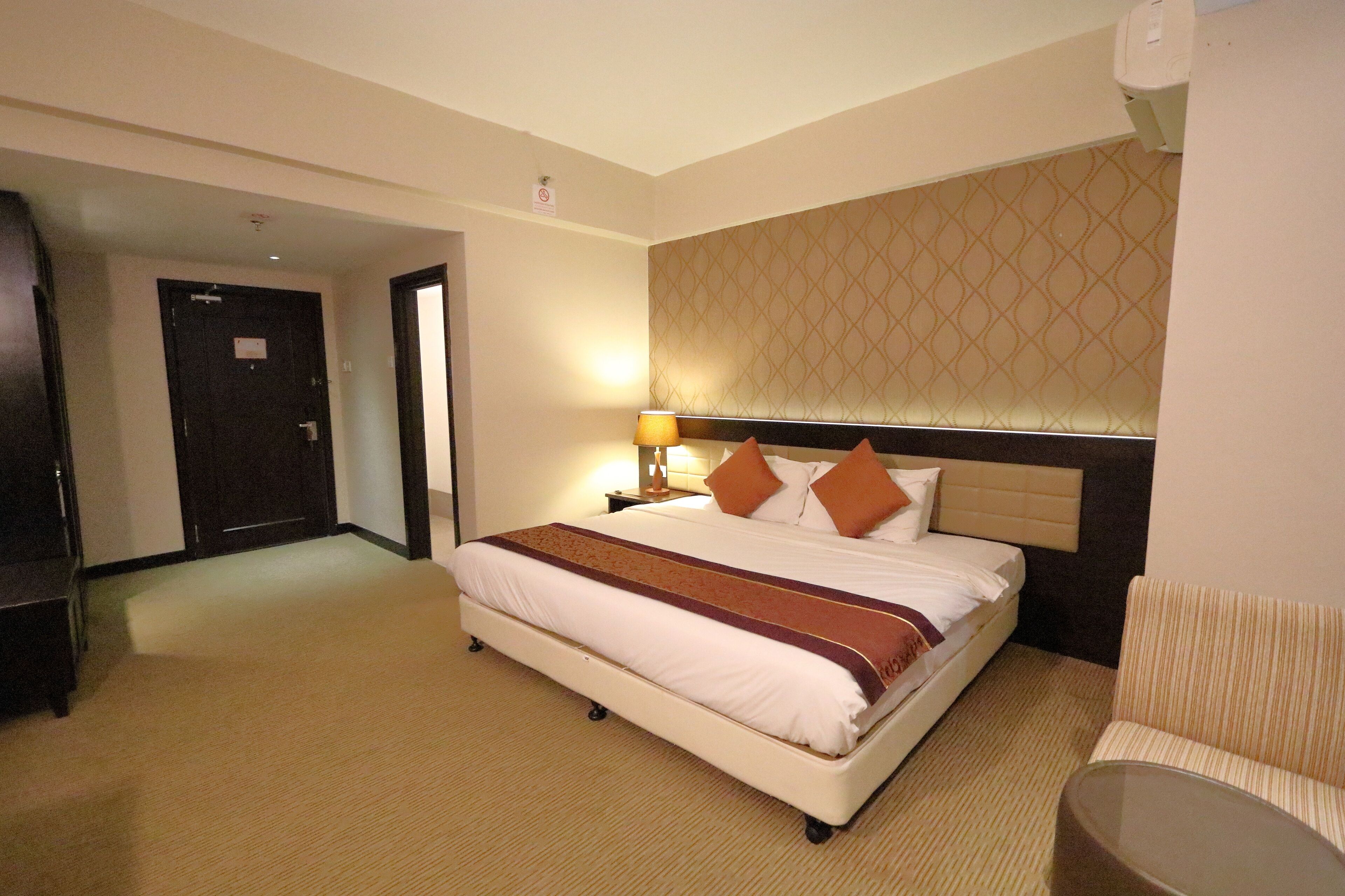 Habitación de hotel con una cama grande, una mesita de noche con lámpara, una silla y suelo cubierto de alfombra.
