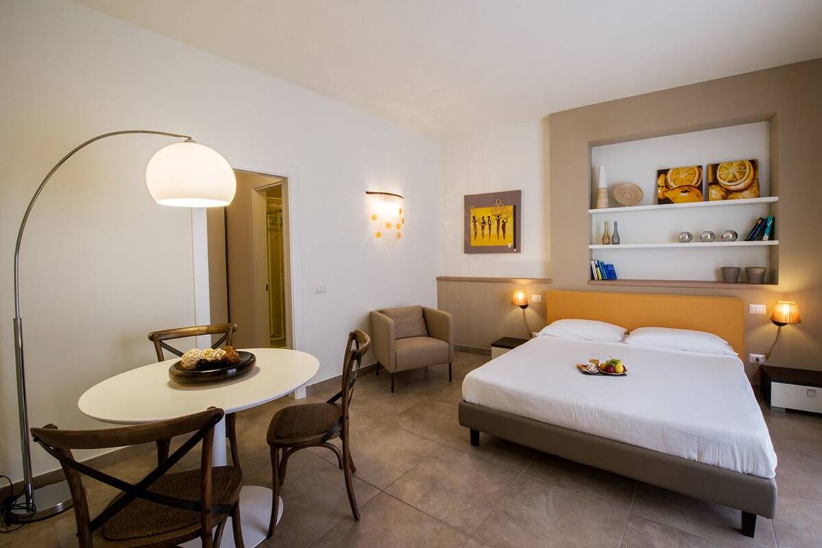 Habitación de hotel con cama, una mesa redonda con sillas, escritorio, lámpara, estante con objetos decorativos y una luz en la pared.
