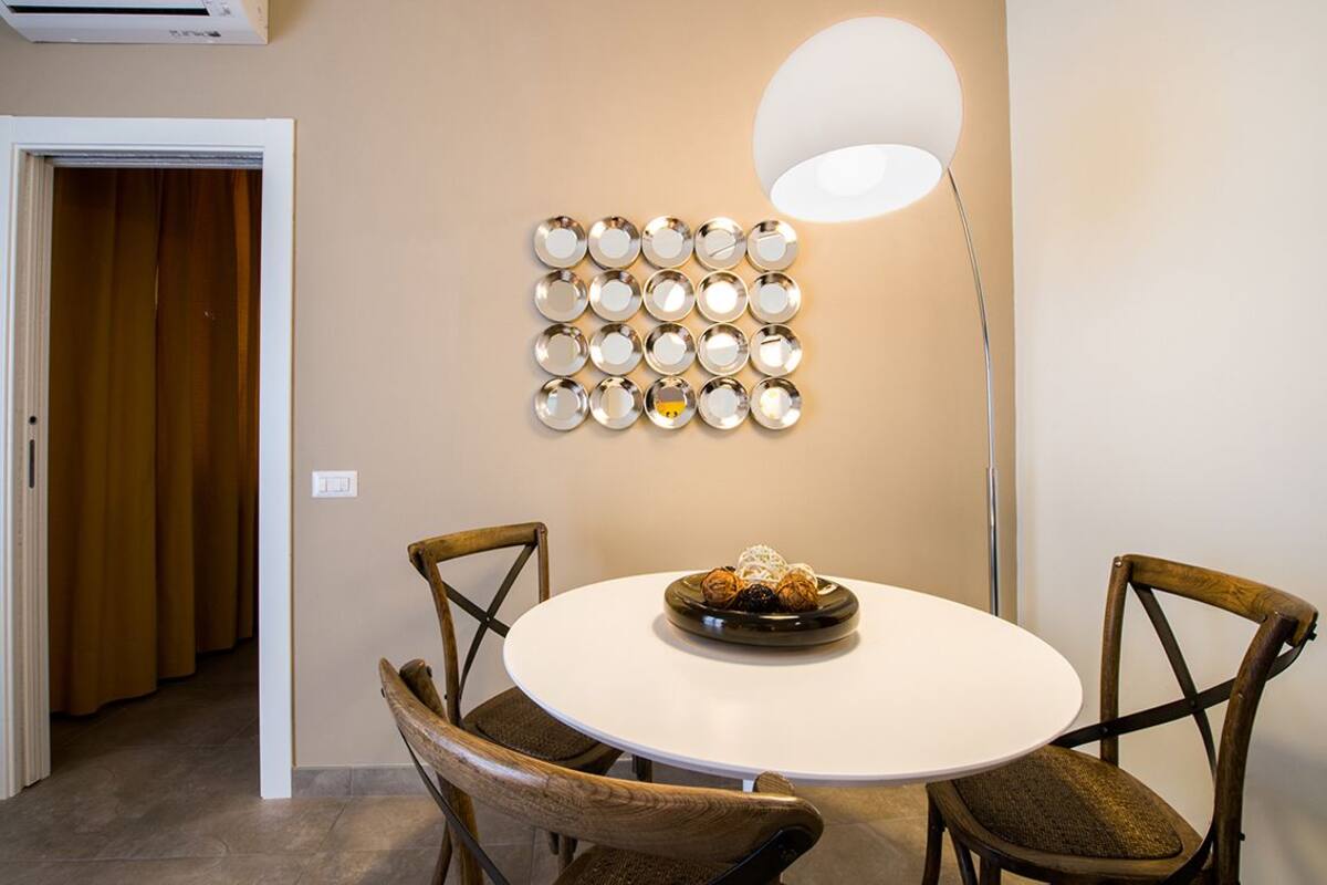 Un comedor con una mesa redonda, dos sillas de madera, una lámpara fijada a la pared y una puerta al fondo.