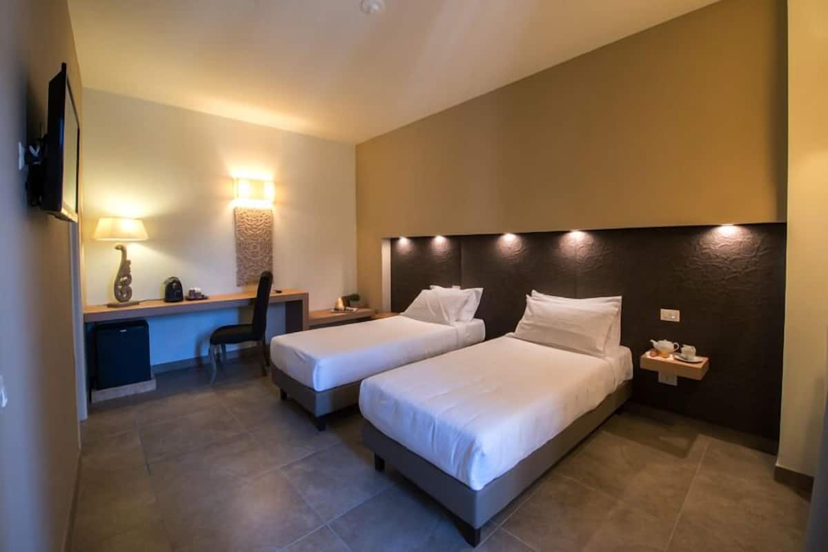 Habitación de hotel con dos camas individuales, un escritorio con silla, televisor montado en la pared y una mesita de noche con taza y platillo.