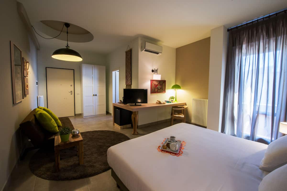 Habitación de hotel con una cama, un escritorio con computadora, televisor, una silla, una mesita con planta y una ventana con cortinas.