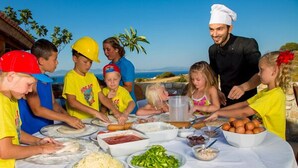 A group of people preparing food outdoors with a chef, various ingredients on the table, and a scenic view in the background. - Aegean View Aqua Resort (Kos)