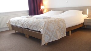 Een tweepersoonsbed met een beige en witte bedspreide, een nachtkastje met een lamp, en een raam met gordijnen.