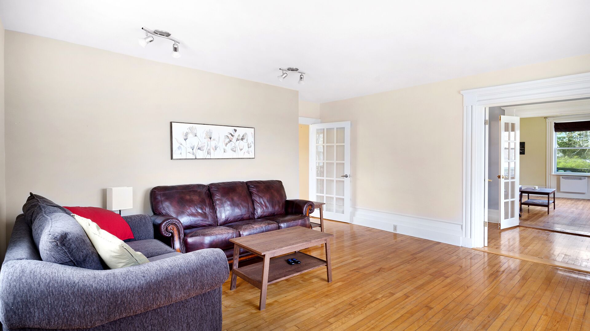A living room with a brown leather sofa, a grey armchair, and a wooden coffee table. There is a framed artwork on the wall and a door leading to another room.