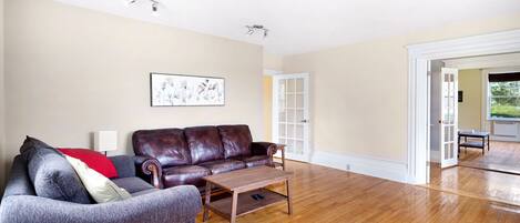 A living room with a brown leather sofa, a grey armchair, and a wooden coffee table. There is a framed artwork on the wall and a door leading to another room.