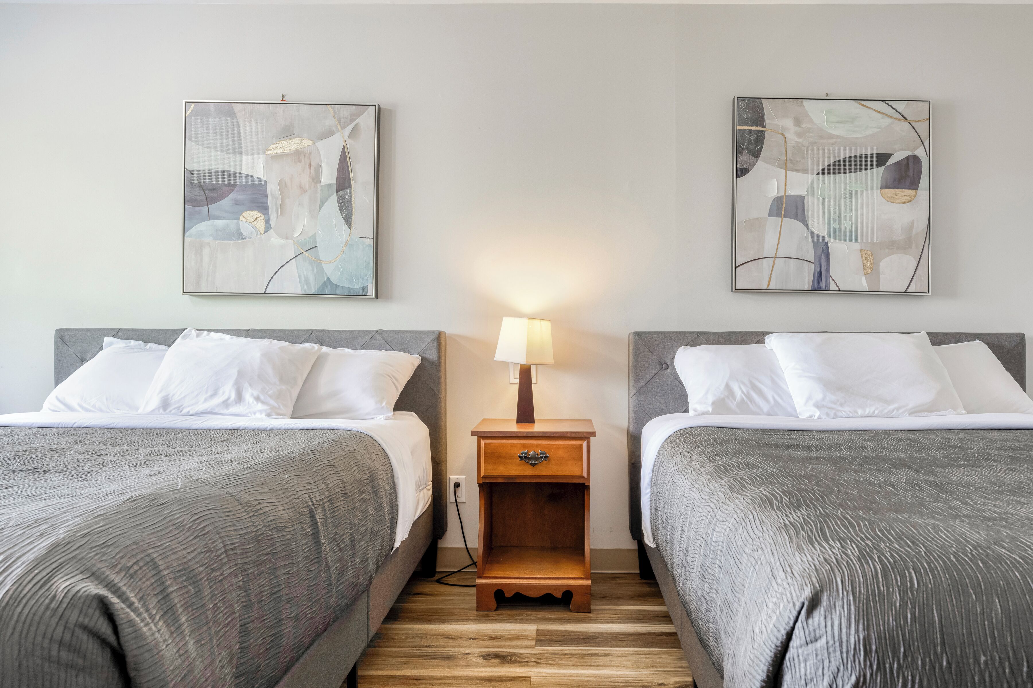 A hotel room with two beds, a nightstand with a lamp, and abstract wall art.