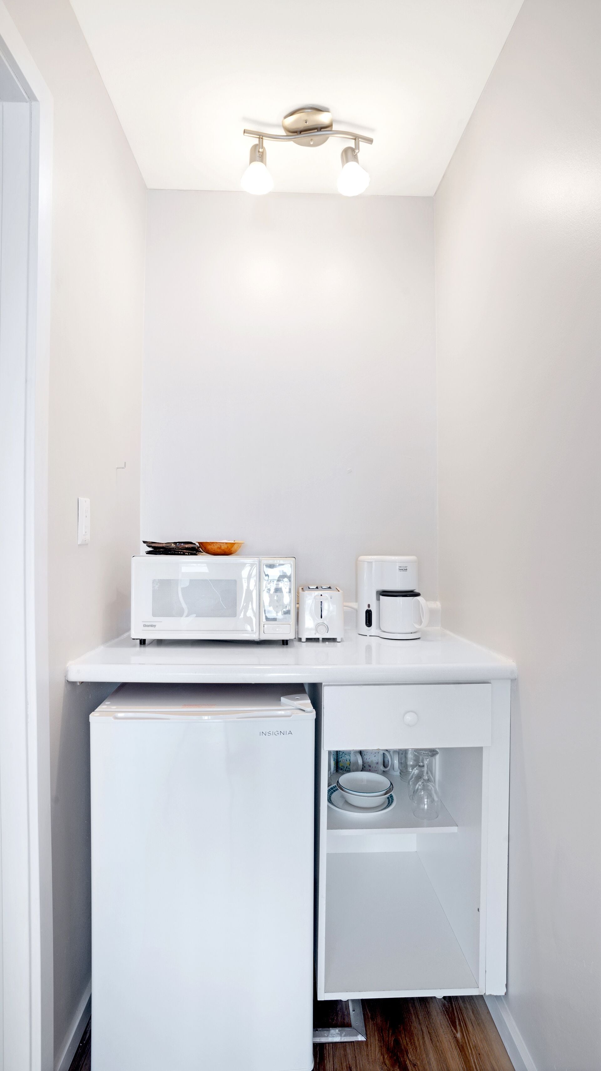 Una kitchenette compacta con microondas, tostadora y cafetera sobre una encimera blanca, un refrigerador pequeño debajo y una lámpara en el techo.