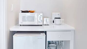 Una kitchenette compacta con microondas, tostadora y cafetera sobre una encimera blanca, un refrigerador pequeño debajo y una lámpara en el techo.