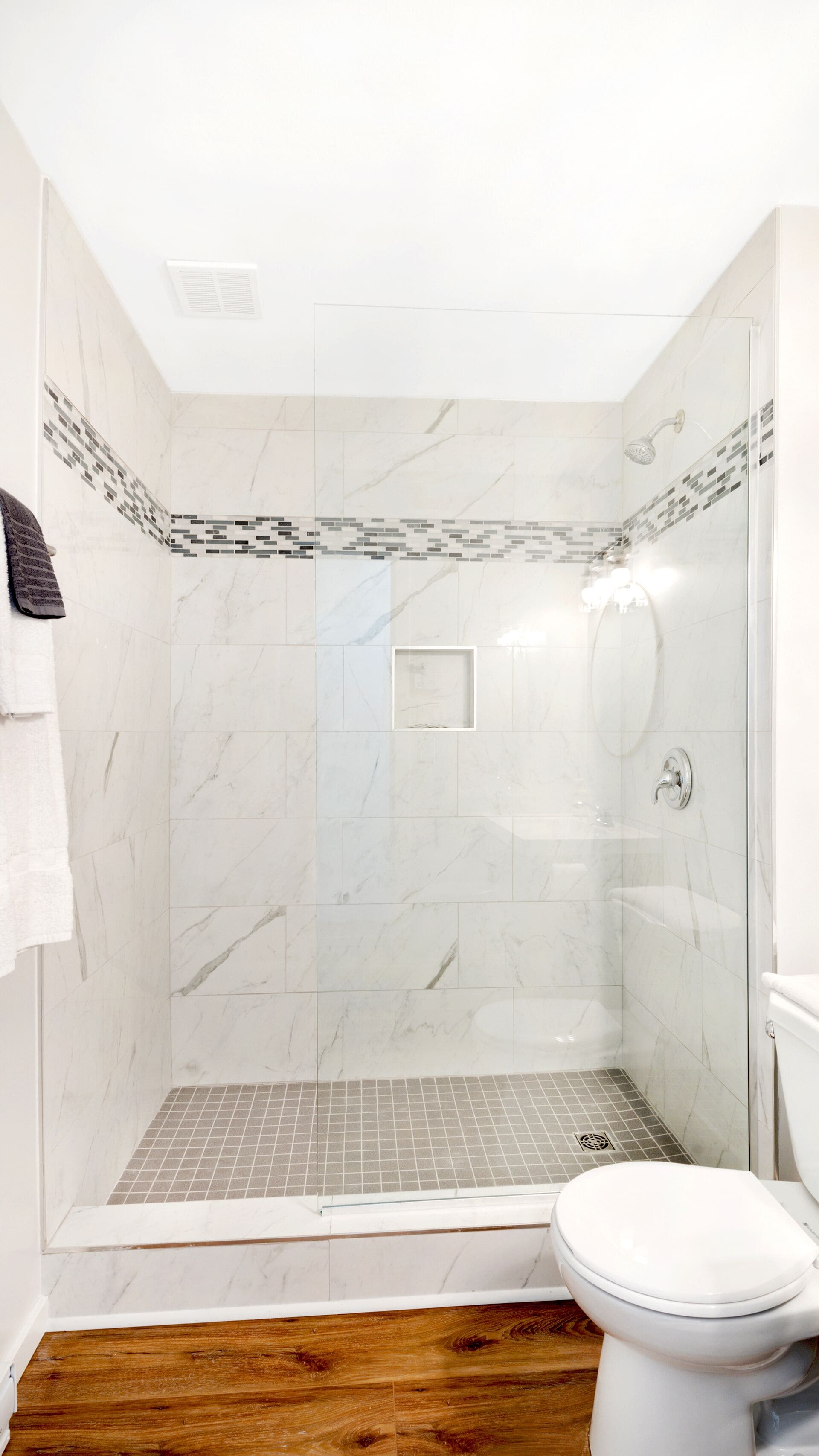 Una ducha con puerta de vidrio, inodoro blanco y suelo de madera.