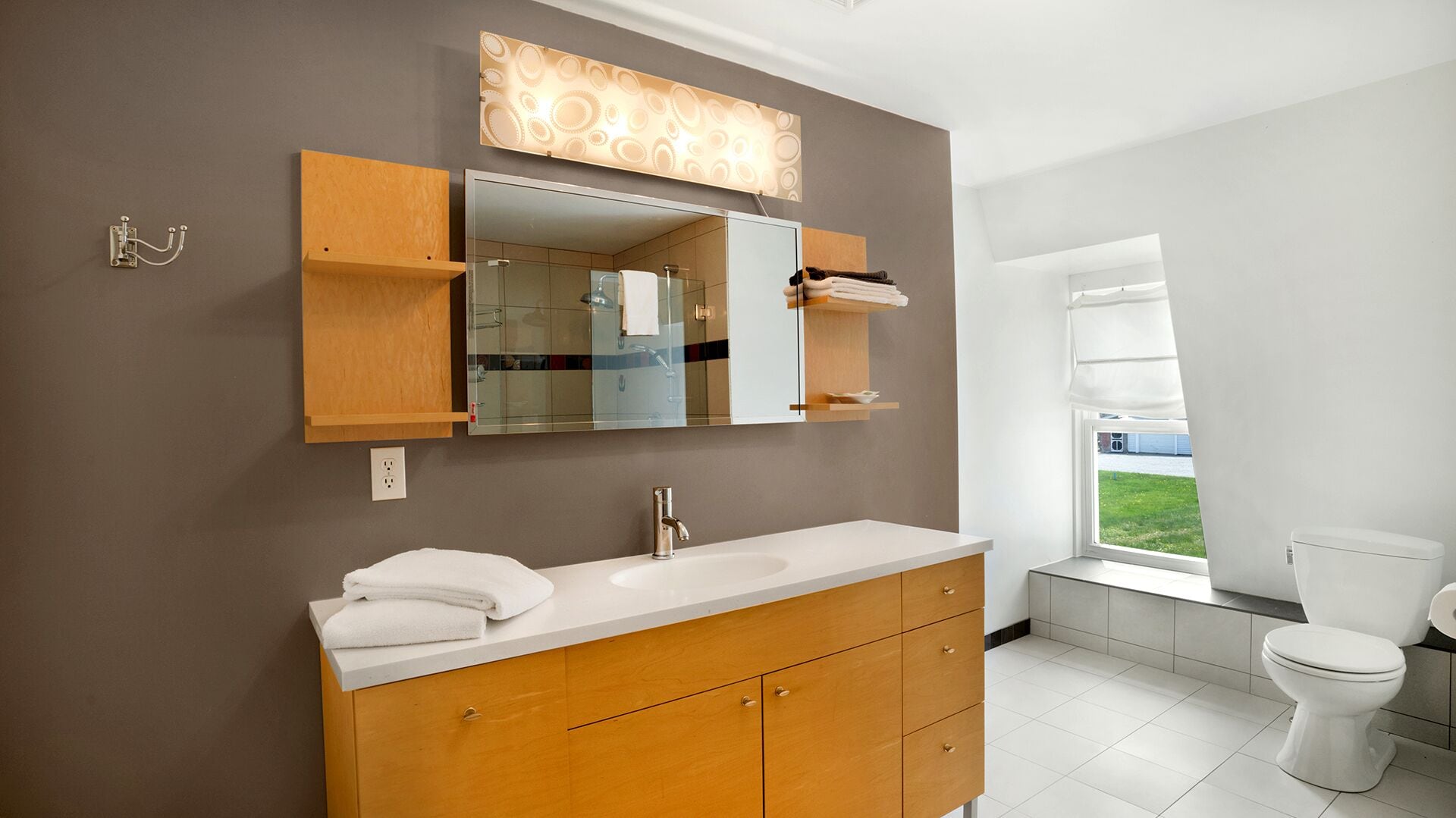 A modern bathroom with a wooden vanity, a large mirror, and a view of the outdoors through a window.