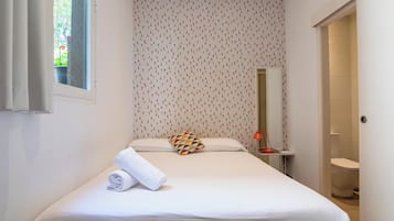 Een net opgemaakt bed met een opgerolde witte handdoek, een gestreept kussen en een klein nachtkastje met een lamp.