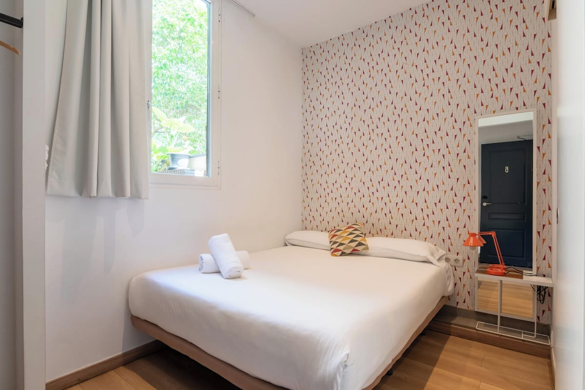 A bedroom with a bed, a window with curtains, a patterned wall, a door, and a nightstand with a lamp.
