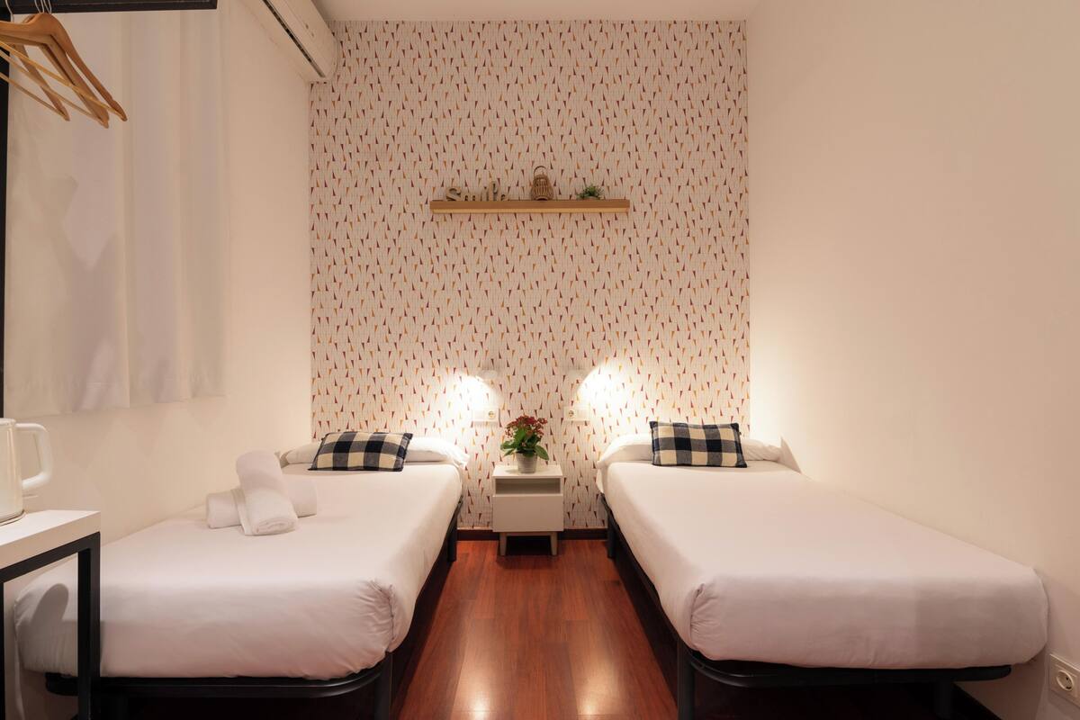 A hotel room with two single beds, a small table with a mug, a shelf with a basket, and a wall with a patterned wallpaper.