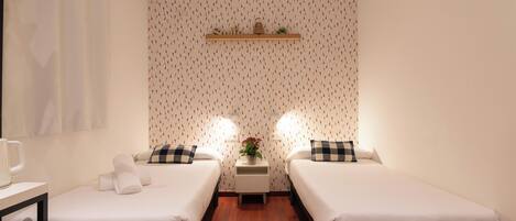 A hotel room with two single beds, a small table with a mug, a shelf with a basket, and a wall with a patterned wallpaper.
