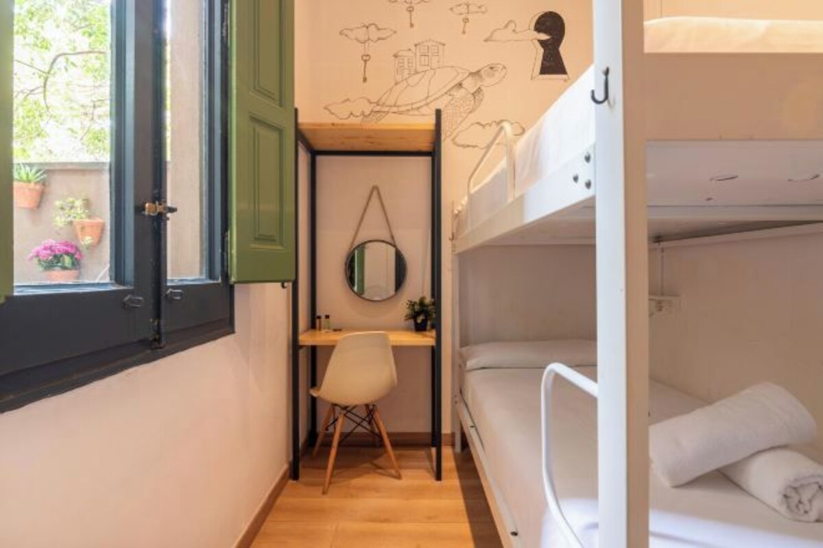 Une chambre de taille modeste, équipée de lits superposés, d’un bureau, d’une chaise et d’une fenêtre donnant sur l’extérieur.