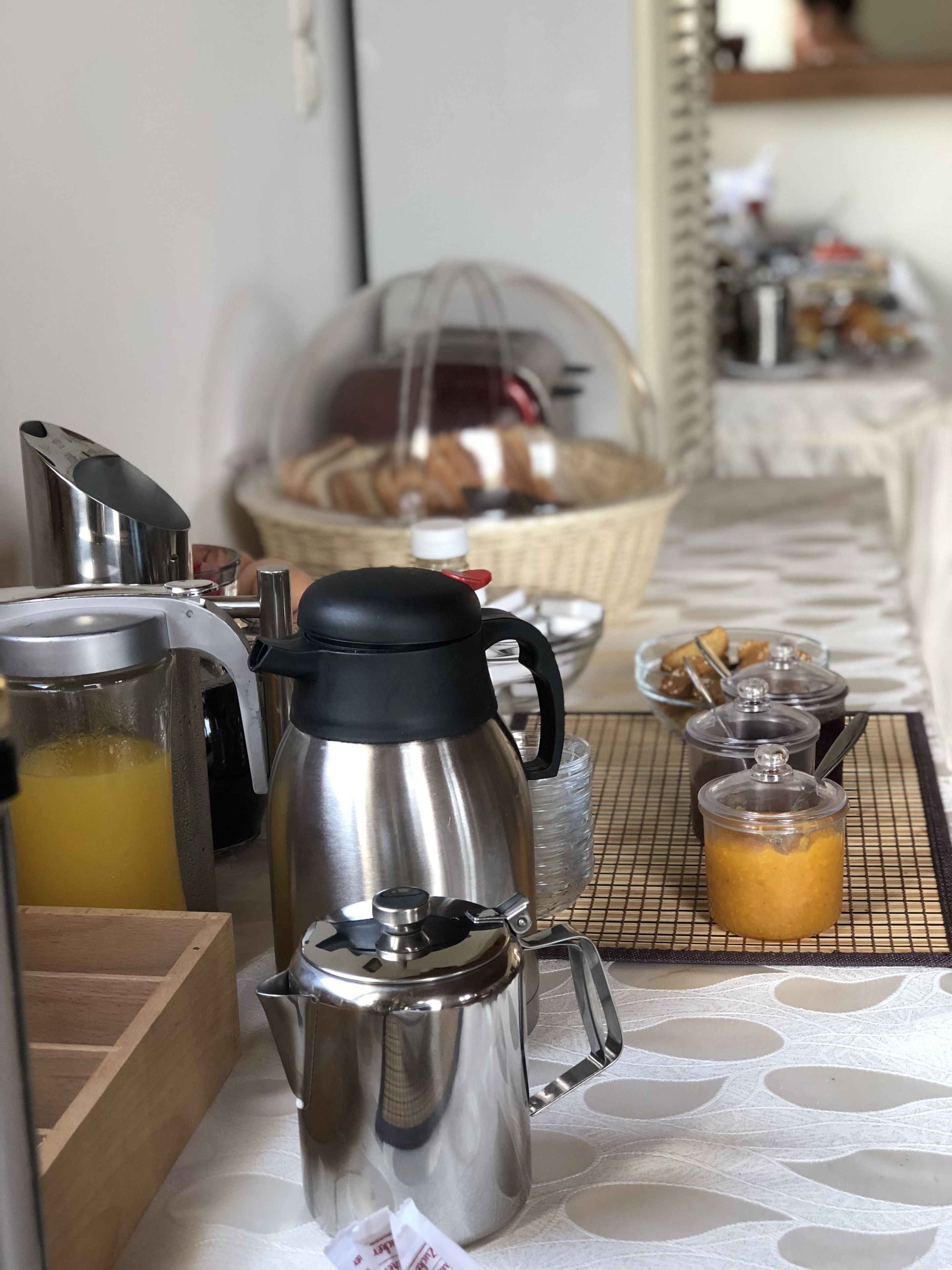 A breakfast setup with a stainless steel coffee pot, a glass carafe with orange juice, and a basket of pastries.