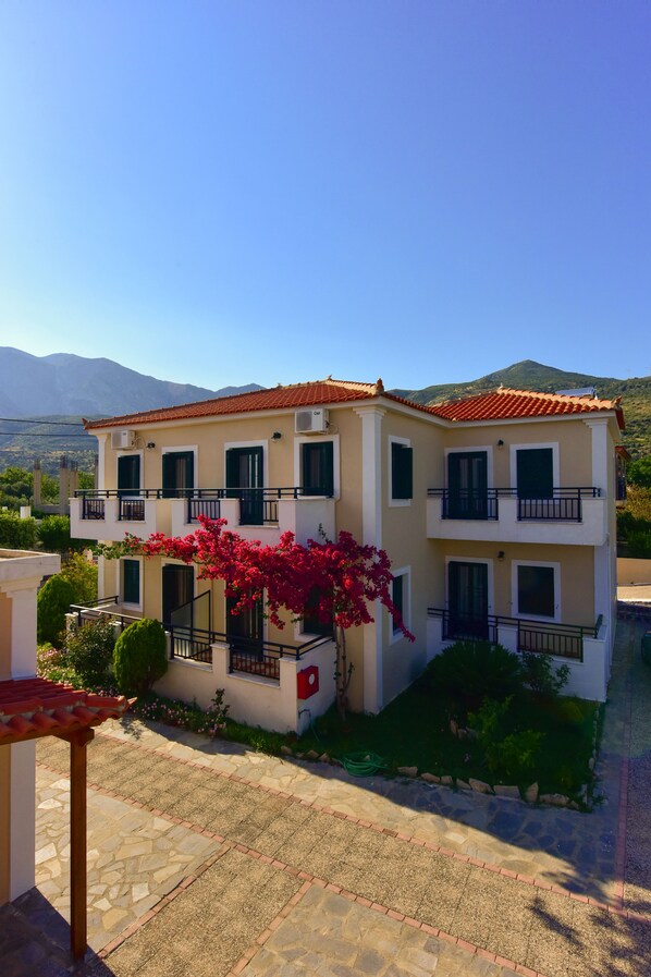 A two-story building with balconies, a tiled roof, and a garden with flowering plants.