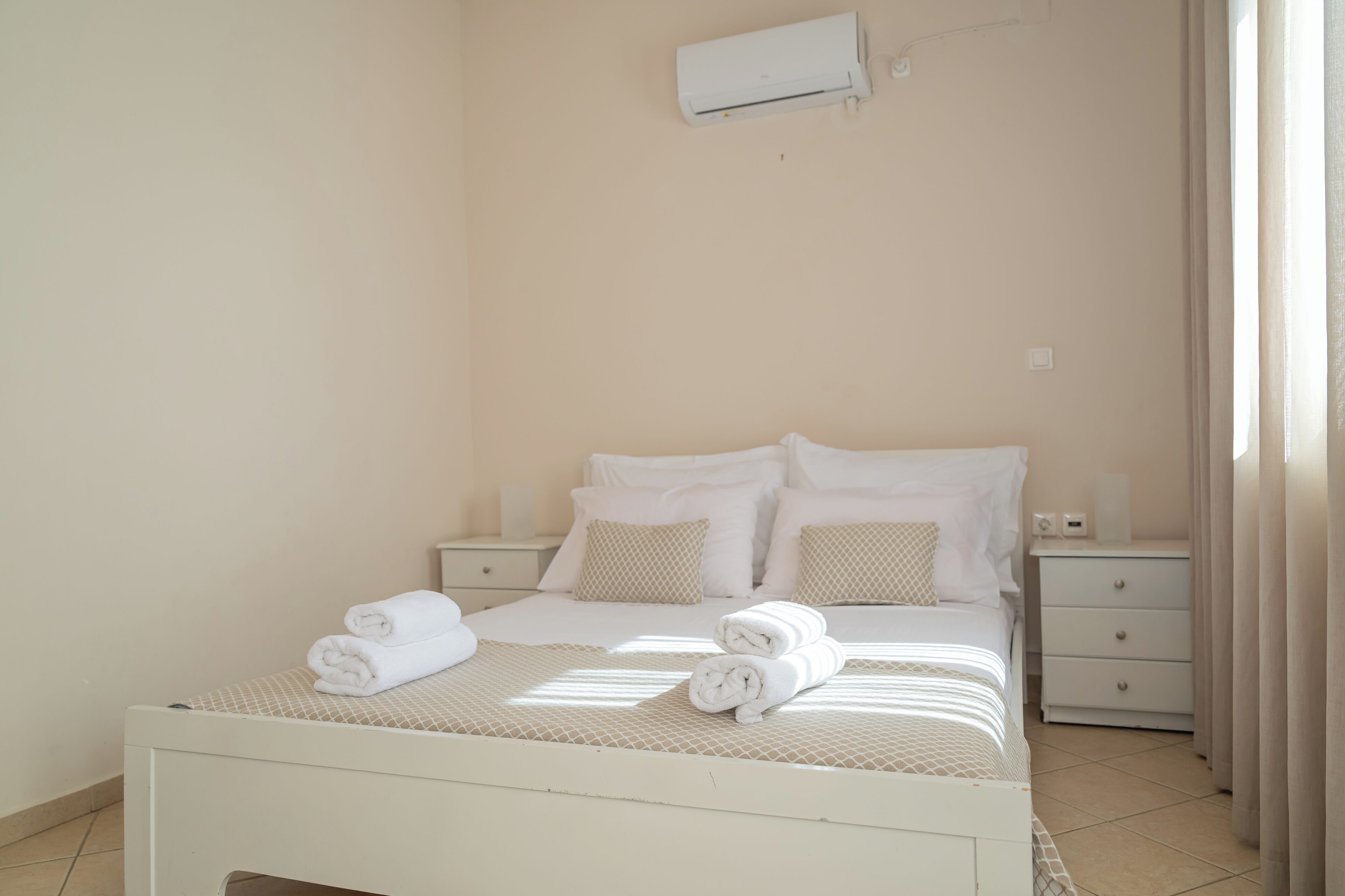 A neatly made bed with white linens and two rolled towels, a bedside table with a lamp, and a wall-mounted air conditioner.