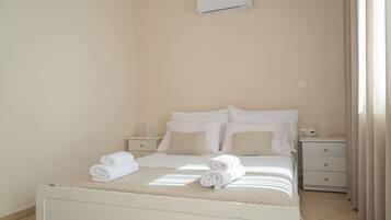 A neatly made bed with white linens and two rolled towels, a bedside table with a lamp, and a wall-mounted air conditioner.