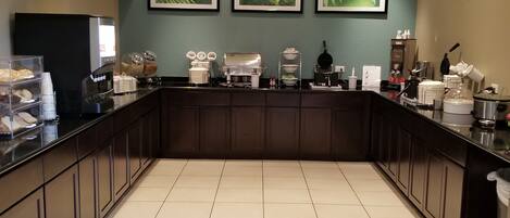 A modern kitchen with a long counter, equipped with coffee machines, a refrigerator, and various kitchen appliances.