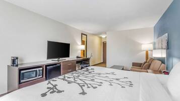 A hotel room with a bed, a television, a desk with a microwave, a chair, and a lamp.