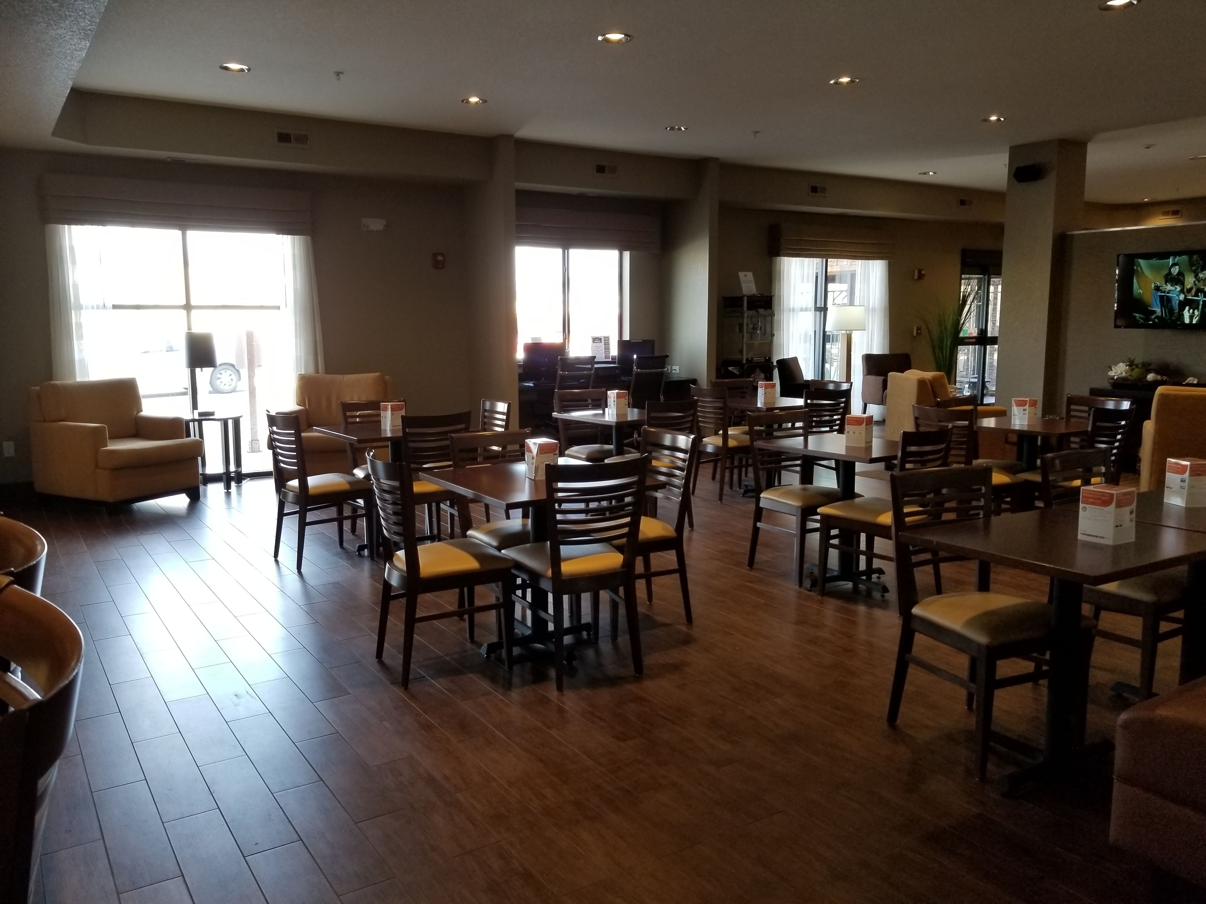 A spacious dining area with multiple tables and chairs, large windows allowing natural light, and a flat-screen TV mounted on the wall.