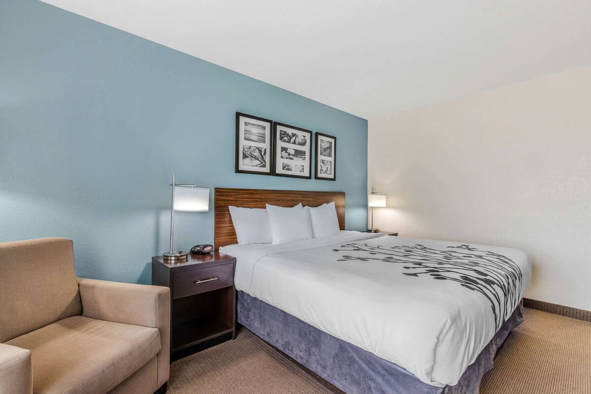 A hotel room with a bed, nightstand, lamp, and a chair.