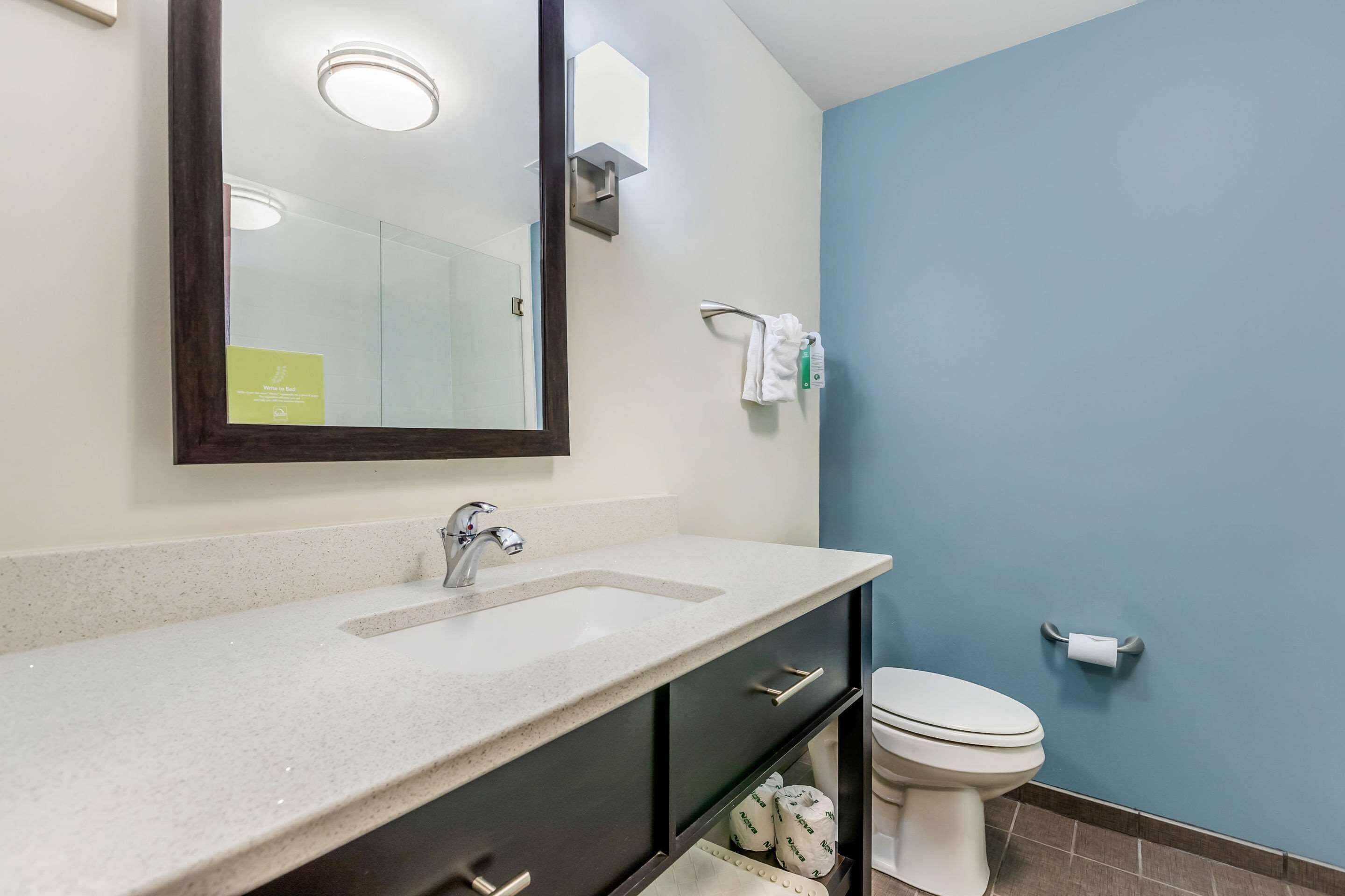 Un baño con lavamanos blanco, muebles oscuros, un espejo, toallero y un inodoro.