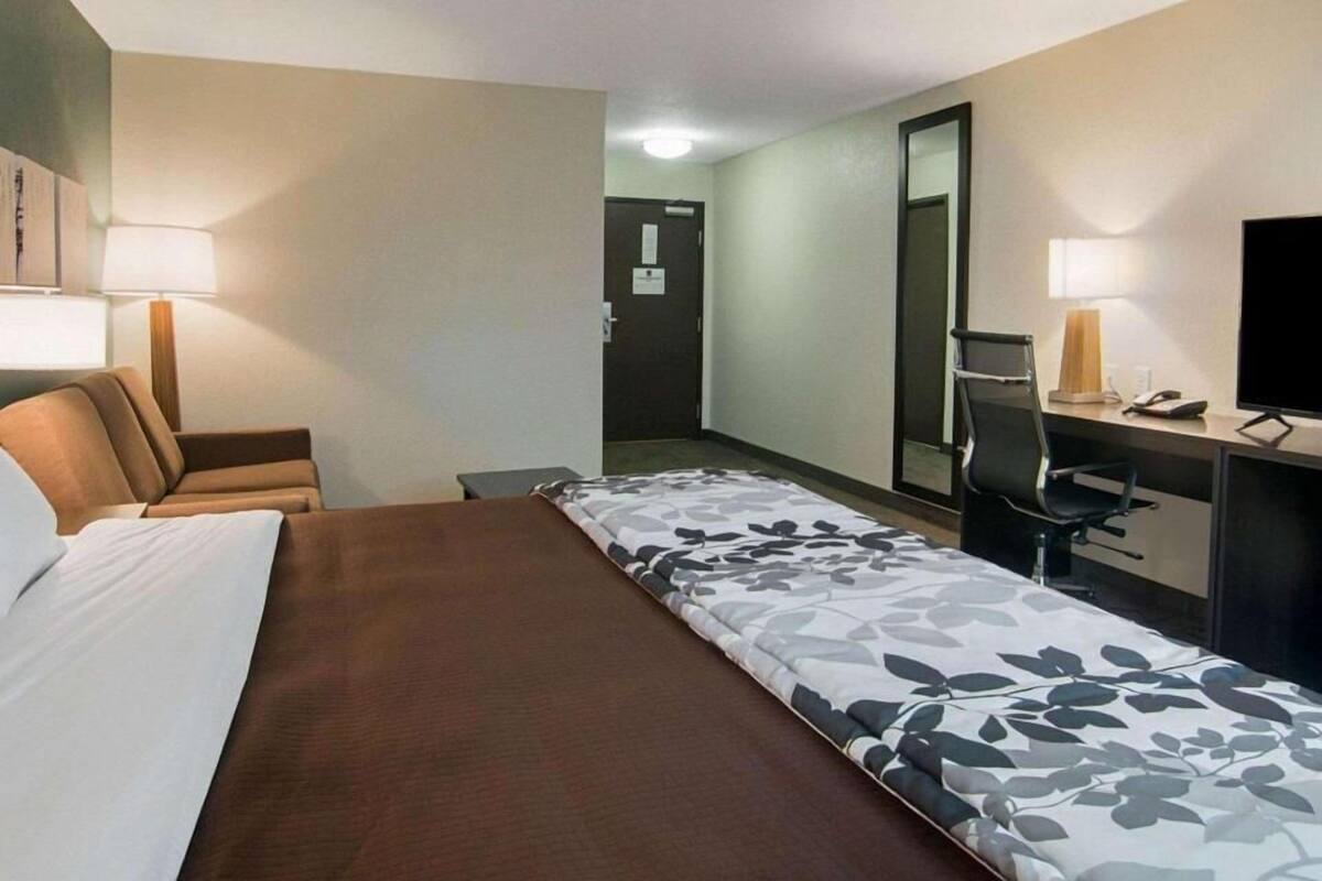 A hotel room with a bed, a desk, a chair, a lamp, a TV, and a door.
