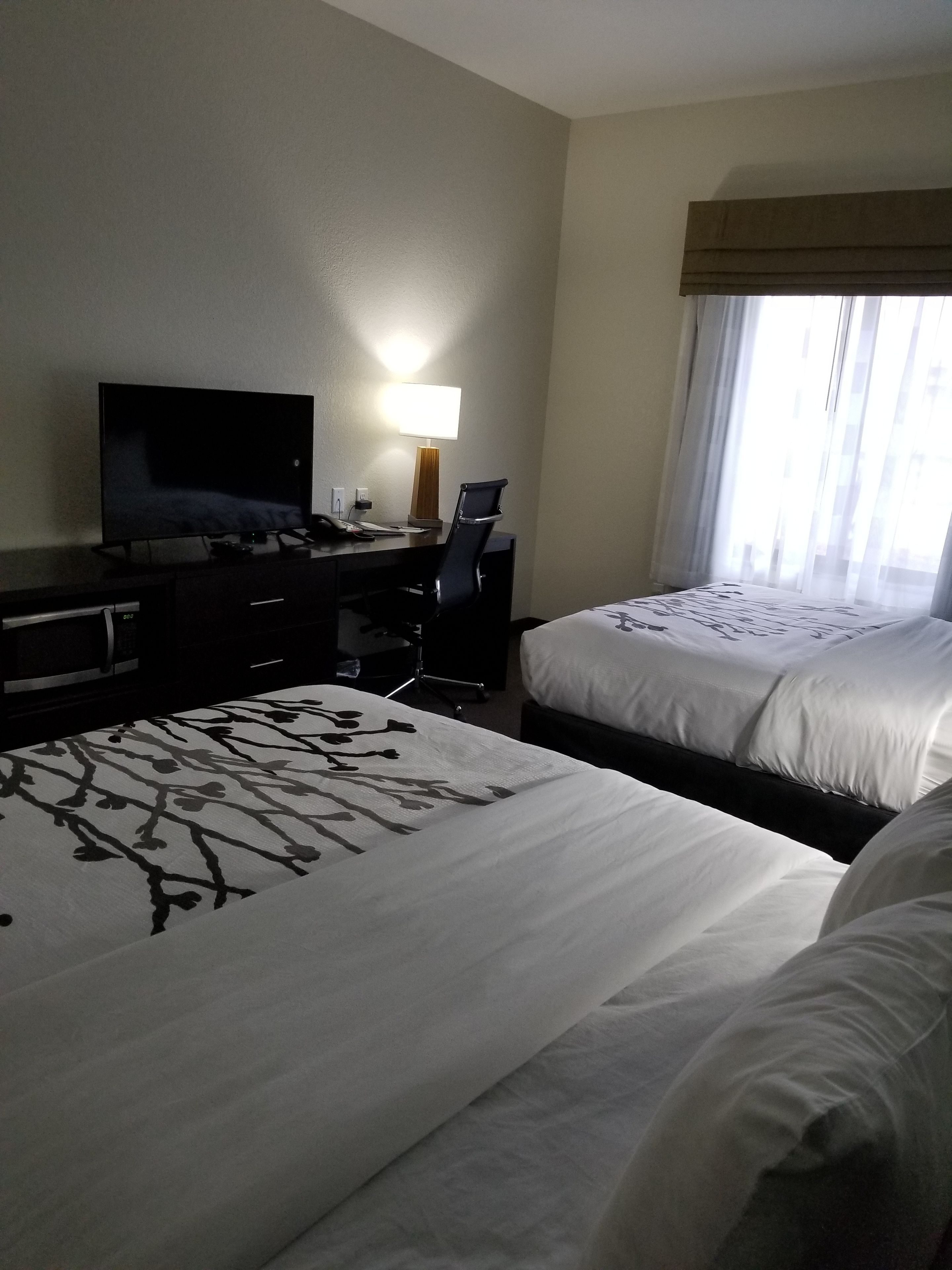 A hotel room with two beds, a desk with a television, a lamp, and a window with curtains.