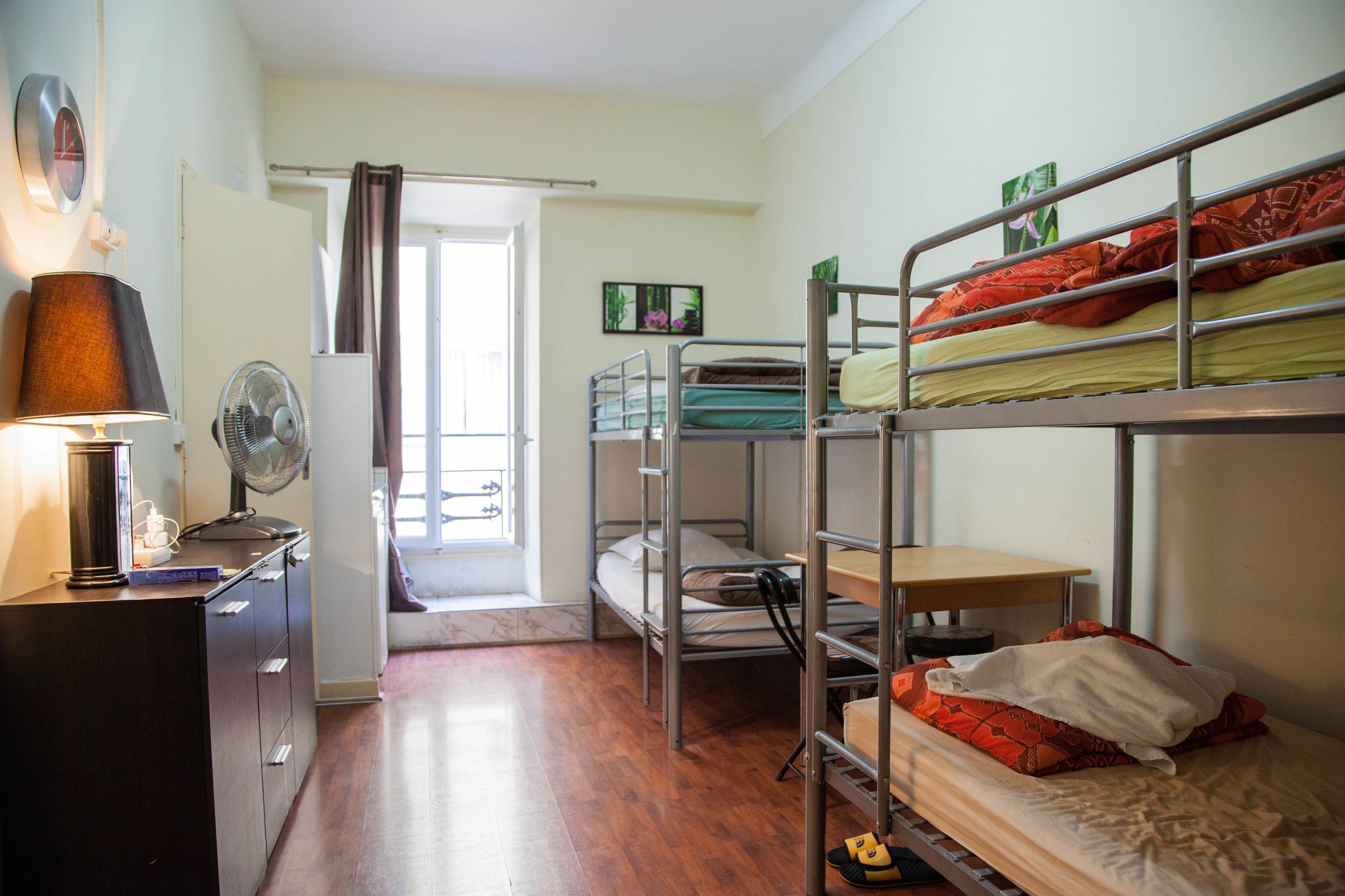 Une chambre de dortoir avec des lits superposés, un bureau, une lampe, un ventilateur et une fenêtre avec un rideau.