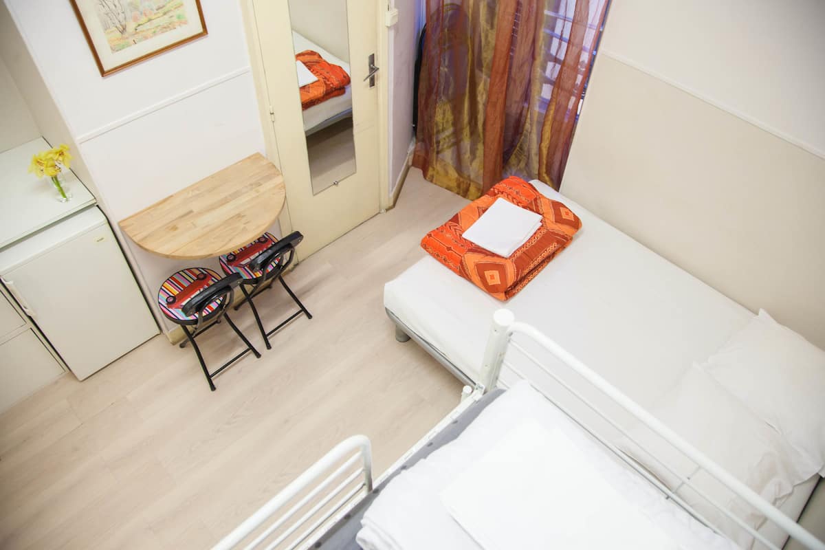 A small, compact room with a bed, a desk, and a chair. There is a door leading to another room.