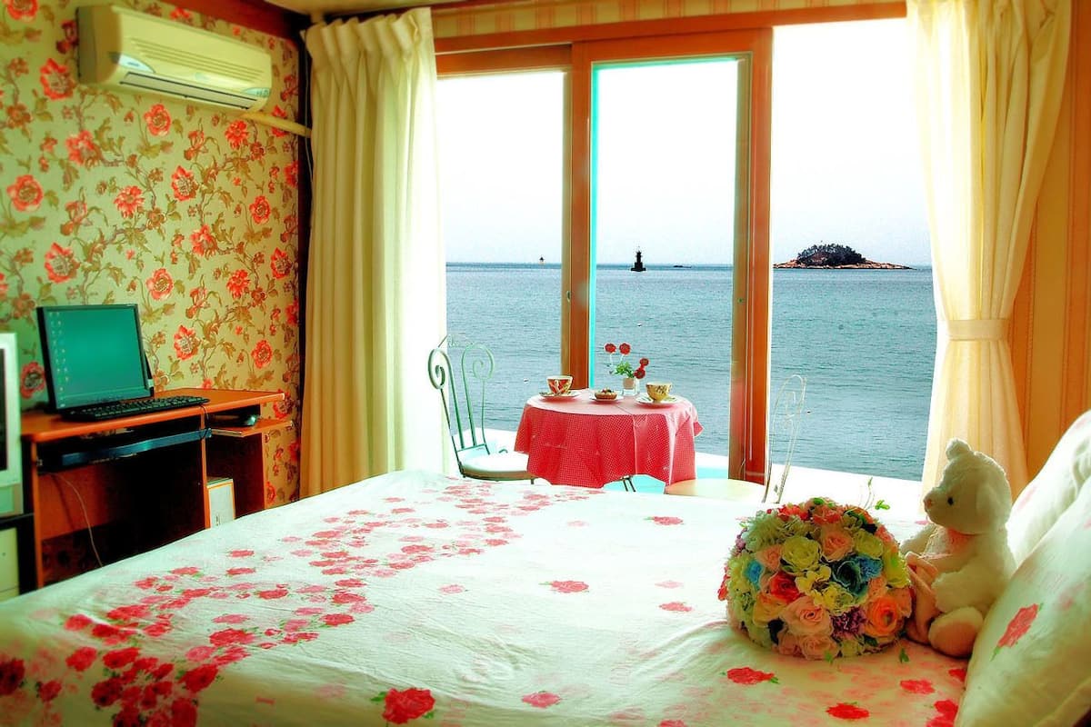 Una habitación con una cama, un escritorio con una computadora portátil, una pequeña mesa con un ramo de flores y vista al mar.