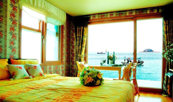 Quarto com cama, mesa com um vaso de flores e vista para barcos na água através de grandes janelas.